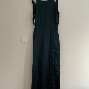 Mörkgrön långklänning - Elegant mörkgrön långklänning i satin med smala axelband och en stilren design. Perfekt för speciella tillfällen med sin vackra fall och glansiga yta. Klänningen har en diskret v-ringning baktill som ger en extra touch av elegans.