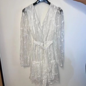 Vit studentklänning - Elegant vit omlottklänning med blommigt mönster och långa ärmar. Klänningen har ett knytband i midjan och en lätt transparent look som ger en romantisk känsla. Perfekt för studenten, eller vårens och sommarens tillfällen. Helt oanvänd