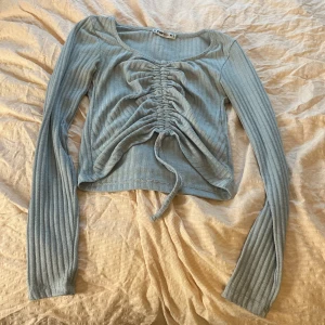 Ljusblå ribbad topp från FB Sister - Säljer en ljusblå ribbad topp från FB Sister med långa ärmar och snygg snörning framtill. Toppen har en croppad passform och är perfekt för en trendig look. Passar bra till både jeans och kjol. Man kan knyta en rosett läst ner på tröjan. 