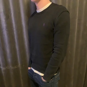 Svart stickad tröja från Ralph Lauren - Snygg svart stickad tröja från Ralph Lauren med en liten broderad logga på bröstet. Tröjan har långa ärmar och ribbstickade muddar vid ärmslut och nederkant. Perfekt för en stilren look.