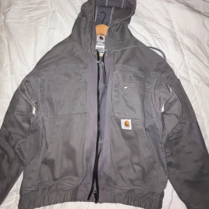 Grey Vintage Recycle jacka från Carhartt, - Säljer en grå jacka från Carhartt med huva och dragkedja. Jackan har flera praktiska fickor och en avslappnad passform. Perfekt för en casual look. 