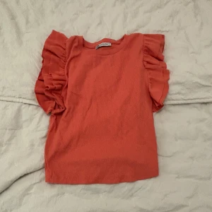 Korallfärgad topp med volang från Zara - Säljer en söt korallfärgad topp från Zara med volangdetaljer längs ärmarna. Toppen är ribbad och har en rund halsringning. Perfekt för att liva upp garderoben med lite färg!