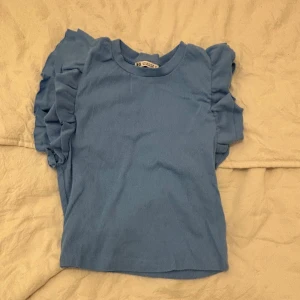 Blå ribbad topp från Zara - Säljer en söt blå ribbad topp från Zara med volangdetaljer på axlarna. Perfekt för en stilren och trendig look. Toppen har en rund halsringning och är tillverkad i ett mjukt material.