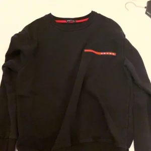 Svart tröja från Prada - Säljer en stilren svart tröja från Prada med en röd och vit detalj på bröstet. Tröjan har långa ärmar och en klassisk rund halsringning. Perfekt för en trendig och avslappnad look.