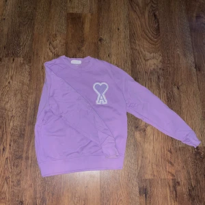 Lila sweatshirt från Ami - Säljer en snygg lila sweatshirt från Ami med ett hjärta och bokstaven A broderat på framsidan. Tröjan har långa ärmar och en rund halsringning. Perfekt för en avslappnad stil.