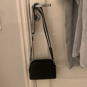 Svart axelväska med randigt band - Snygg svart axelväska med ett brett, justerbart randigt band i svart och vitt. Väskan har en dragkedja upptill och är perfekt för att bära dina nödvändigheter. Den kompakta designen gör den lätt att bära med sig överallt. Bandet är avtagbart och kedjorna är lite slitna men annars i fint skick❤️