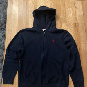 Mörkblå hoodie från Polo Assn - Säljer en snygg mörkblå hoodie från Us polo Assn med dragkedja och huva. Den har en röd broderad logga på bröstet och praktiska fickor framtill. Perfekt för en avslappnad stil.