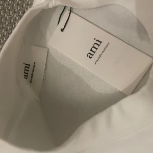 Vit t-shirt från Ami - Säljer en stilren vit t-shirt från Ami med ett rött hjärta och bokstaven A broderat på bröstet. Perfekt för en enkel och snygg look. T-shirten har en normal passform och är kortärmad.