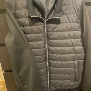 Svart quiltad jacka - Snygg svart quiltad jacka med dragkedja och ribbad krage. Perfekt för kyligare dagar med sin stilrena design och bekväma passform. Jackan har långa ärmar och praktiska fickor framtill.