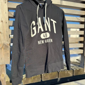 Svart hoodie från GANT - Säljer en stilren svart hoodie från GANT med tryck '49 New Haven' på framsidan. Tröjan har långa ärmar och en klassisk känguruficka. Perfekt för en avslappnad stil. 🖤