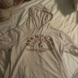 Beige Stanford hoodie - Säljer en beige Stanford hoodie med tryck framtill. Den har en klassisk känguruficka och en bekväm huva med dragsko. Perfekt för en avslappnad stil eller för att mysa i hemma. Passar bra till både jeans och mjukisbyxor.