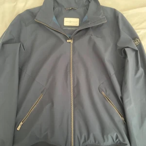 Blå vindjacka från Sail Racing - Säljer en snygg blå vindjacka från Sail Racing i storlek L. Jackan har en dragkedja framtill och två praktiska fickor med dragkedjor. Perfekt för blåsiga dagar och har en stilren design med logga på ärmen. Pris kan diskuteras