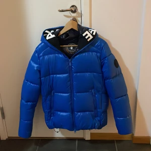 Blå pufferjacka från Save the Duck - Säljer en snygg blå pufferjacka från Save the Duck i mycket bra skick, Edgard Padded Puffer som har orginalpriset på 3700. Den är inte använd utomhus någon gång. Den har en dragkedja framtill och en huva för extra värme. Perfekt för kalla vinterdagar! 