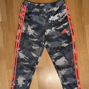 Camouflage träningsbyxor från Adidas - Säljer ett par snygga träningsbyxor från Adidas i camouflagemönster. Byxorna har de klassiska tre ränderna i rött längs sidorna och en elastisk midja för bästa passform. Perfekta för träning eller chill. De är i mycket bra skick och redo för nya äventyr!