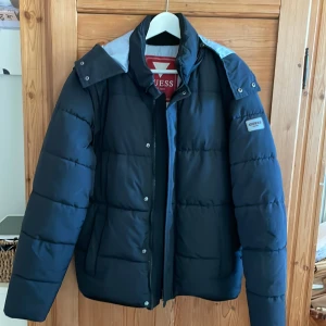 Svart pufferjacka från Guess - Säljer en snygg svart pufferjacka från Guess i mycket bra skick. Jackan har en praktisk huva och dragkedja framtill. Perfekt för kalla vinterdagar med sin varma och quiltade design. Storlek M. Kan användas som väst då ärmarna kan tas av. 