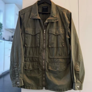 Olivgrön jacka - Säljer en olivgrön jacka i bra skick från lager 157. Den har en dragkedja framtill och flera praktiska fickor. Perfekt för höst och vår med sin avslappnade stil och justerbara midja. Frakt ingår.