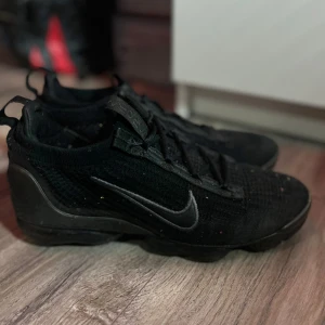 Svarta Nike Vapormax sneakers - Säljer ett par svarta Nike Vapormax sneakers i bra skick. De har en stilren design med snörning och är perfekta för både träning och vardagsbruk. Skorna är gjorda i syntetmaterial och har en bekväm passform. Perfekta för dig som vill ha både stil och komfort! En sula har lite mindre luft än den andra men det går att fylla på. Priset är inte hugget i stel men kan diskuteras!!!( Ena skorn har hål i bubbelsulan)