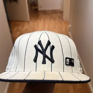 Säljer en snygg vit keps med blå ränder från New Era. Den har en broderad logga framtill och är i mycket bra skick. Perfekt för sommarens alla äventyr eller som en stilren accessoar till vardagsoutfiten.