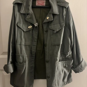 Grön jacka från Levi Strauss & Co - Säljer en grön jacka från Levi Strauss & Co i storlek M. Jackan har coola pins som man även kan ta av. Den har knappar framtill och två bröstfickor med lock. Perfekt för höst och vår!