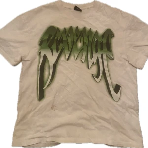 Vit t-shirt med tryck från Revenge - Säljer en cool vit t-shirt från Revenge med ett stort grönt tryck på framsidan och en häftig dödskalle på baksidan. Perfekt för en avslappnad stil eller en konsertkväll. T-shirten är i bomull och har en normal passform. Grymt snygg och unik!