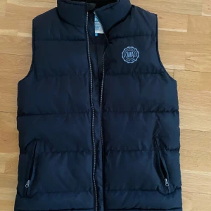 Svart dunväst från WA - Säljer en svart dunväst från WA i mycket bra skick. Västen har en dragkedja framtill och två fickor med dragkedjor. Perfekt för höst och vår, med en stilren design och ett broderat märke på bröstet. Superbekväm och lätt att matcha med andra kläder!