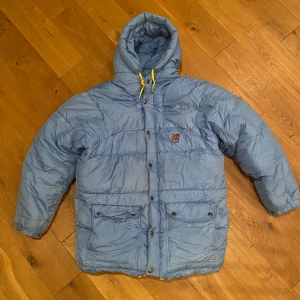 Ljusblå fjällräven  - Äldre Fjällrävenjacka säljes. Färg: Ljusblå . Storlek: Det står S/M men jag tycker att den är mer som medium  Spår av användning finns. Bland annat en mindre ljus fläck på ryggen se bild 2. Se även bild 4 där locket på fickan inte är helt slät. Knapparna fungerar utan anmärkning. Vid ärmslut och under armar finns tecken på användning. En tvätt rekommenderas. Inga skador. En supervarm och väldigt bra jacka.