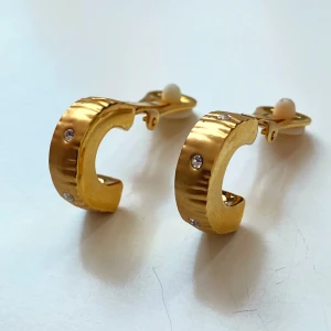Guldörhängen med stenar clips  - Snygga guldörhängen med små gnistrande stenar. De har en rundad form och är perfekta för att ge en touch av glamour till vilken outfit som helst. Passar både till vardag och fest! Saknar en rhinestone, i övrigt mycket bra skick. Okänd metall 