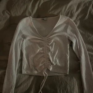 Vit ribbad topp med knytning - Säljer en superfin vit ribbad topp från Zara. Den har en snygg v-ringning och långa ärmar. Framtill finns en trendig knytning som ger en cool look. Perfekt för både vardag och fest!