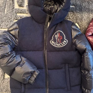 Mörkblå dunjacka från Moncler - Säljer en snygg mörkblå dunjacka från Moncler. Jackan har en quiltad design med glansiga ärmar och en broderad Moncler-logga på bröstet. Den är perfekt för kalla dagar med sin varma vaddering och dragkedja framtill. Huvan ger extra skydd mot väder och vind. Passar perfekt för vintern!