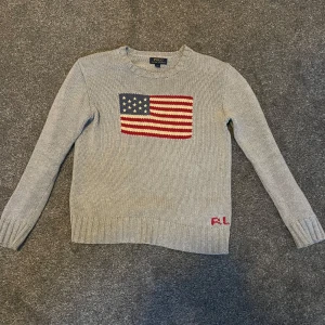 Grå stickad tröja från Ralph Lauren - Säljer en snygg grå stickad tröja från Ralph Lauren med en cool amerikansk flagga på framsidan. Tröjan har långa ärmar och är i mycket bra skick. Perfekt för höst och vinter. Passar både till vardags och lite finare tillfällen. Hund i hemmet! 14-16 år 