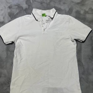 Vit pikétröja från Hugo Boss - Säljer en stilren vit pikétröja från Hugo Boss. Den har en klassisk krage med svarta detaljer och knappar framtill. Tröjan är kortärmad och har en diskret broderad logga på bröstet samt en större logga på ryggen. Perfekt för en avslappnad men snygg look!