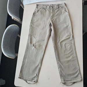 Byxor från Carhartt - Säljer ett par byxor från Carhartt i bra skick. De har en avslappnad passform med flera praktiska fickor och en sliten look med fransar vid fållen. Perfekta för en casual stil eller arbete utomhus.
