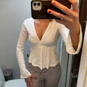 Vit topp ifrån Ginatricot  - Säljer en superfin vit topp från Ginatricot i mycket bra skick. Den har långa ärmar och en v-ringad design med knytband i midjan. Perfekt för både vardag och fest! Passar bra till jeans eller kjol!