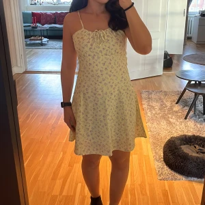 Gul blommig sommarklänning - Söt gul klänning med blommigt mönster i rosa och grönt. Den har smala justerbara axelband och en knytning framtill. Perfekt för sommardagar och midsommarfirande. Klänningen är kort och har en bekväm passform med elastisk rygg för extra komfort. Den är även dubbel fodrad så den är inte genomskinlig ✨