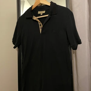 Svart Burberry pikétröja med rutiga detaljer - Säljer en stilren svart burberry pikétröja med en klassisk krage och korta armar i väldigt bra skick, enbart använd 2-3 gånger. Storlek S (passar även M) Den har en snygg och lyxig detalj med ett rutigt mönster vid knäppningen. Perfekt till både vardags och fest, inköpt för 4500kr! 🖤