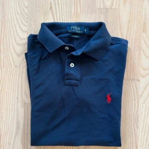 Mörkblå pikétröja från Ralph Lauren - Säljer en klassisk mörkblå pikétröja från Ralph Lauren i storlek S. Tröjan har en röd broderad logga på bröstet och knappar framtill. Perfekt till vardagen. Hör av dig för fler frågor eller funderingar!