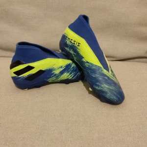 Adidas Nemeziz fotbollsskor - Säljer ett par Adidas Nemeziz fotbollsskor i blått och gult. De har en cool design med mönster som ser ut som penseldrag. Skorna är i mycket bra skick och perfekta för fotbollsspel på gräs. De har en tight passform och är utan snörning för en smidig känsla på planen.