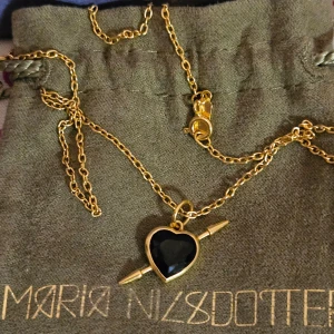 Guldhalsband med svart hjärthänge - Lägger upp en intresskoll. Snyggt guldhalsband från Maria Nilsdotter med ett svart hjärthänge. Hänget har en pil som går igenom hjärtat, vilket ger en cool och unik look. Perfekt för att piffa upp vilken outfit som helst!