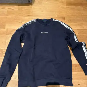 Säljer en snygg mörkblå sweatshirt från Champion i storlek 2X-Large i junior stl .Tröjan har långa ärmar och coola logoband längs ärmarna. Perfekt för en avslappnad stil eller träning. Den är i bra skick och redo för nya äventyr!