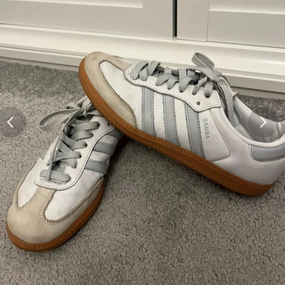 Säljer ett par klassiska Adidas Samba sneakers i vitt och grått. De har en snygg retrostil med bruna sulor och de ikoniska tre ränderna på sidan. Perfekta för både vardag och träning. Skorna är i bra skick med lite slitage på tån. Säljer då de va lite små för mig💕. Skor.