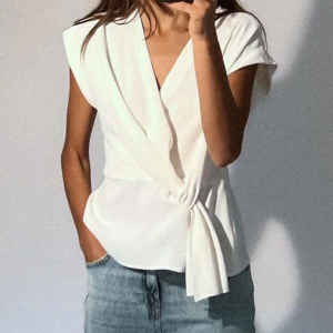 Vit omlottopp från Zara - Säljer en stilren vit omlottopp från Zara. Toppen har en elegant knytning i sidan och är ärmlös, vilket gör den perfekt för både vardag och fest. Materialet är mjukt och bekvämt, vilket ger en fin passform. Passar perfekt till jeans eller kjol för en chic look.