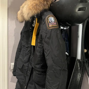 Svart vinterjacka från Parajumpers - Säljer en svart Parajumpers vinterjacka i XXS. Den har en fluffig pälsfodrad huva och är perfekt för kalla dagar. Jackan har flera praktiska fickor och en snygg patch på ärmen. Den är i mycket bra skick och redo för vintern! ❄️