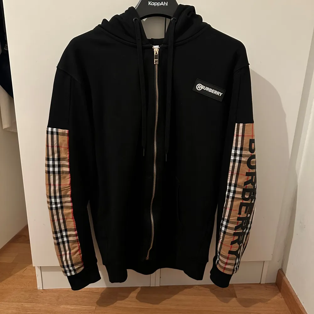 Säljer en snygg svart hoodie från Burberry med dragkedja och klassiska rutiga detaljer på ärmarna. Perfekt för höst och vintern! Den har en bekväm passform och är i mycket bra skick. Perfekt för dig som vill ha en stilren och lyxig look.. Tröjor & Koftor.