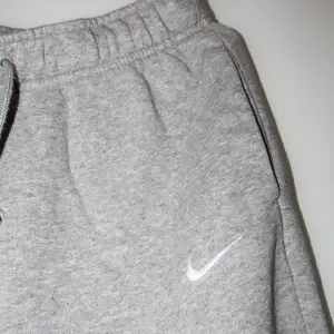 Säljer ett par grå mjukisbyxor från Nike i mycket bra skick. De har en bekväm passform med resår och snörning i midjan. Perfekta för en avslappnad dag hemma eller lätt träning. Nike-loggan är broderad på sidan. Supermjuka och sköna!