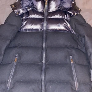 Svart pufferjacka med glansiga detaljer - Säljer en snygg svart pufferjacka med glansiga detaljer och dragkedja framtill. Jackan har en hög krage och avtagbar huva, perfekt för kalla vinterdagar. Den är i mycket bra skick och har praktiska fickor med dragkedjor. Perfekt för både vardag och festliga tillfällen!