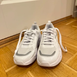 Vita sneakers från Puma - Säljer ett par vita Puma sneakers i nytt skick. De har en stilren design med snörning och en bekväm sula. Perfekta för vardagsbruk eller träning. Skorna är gjorda i syntetmaterial med meshdetaljer för extra andningsförmåga.