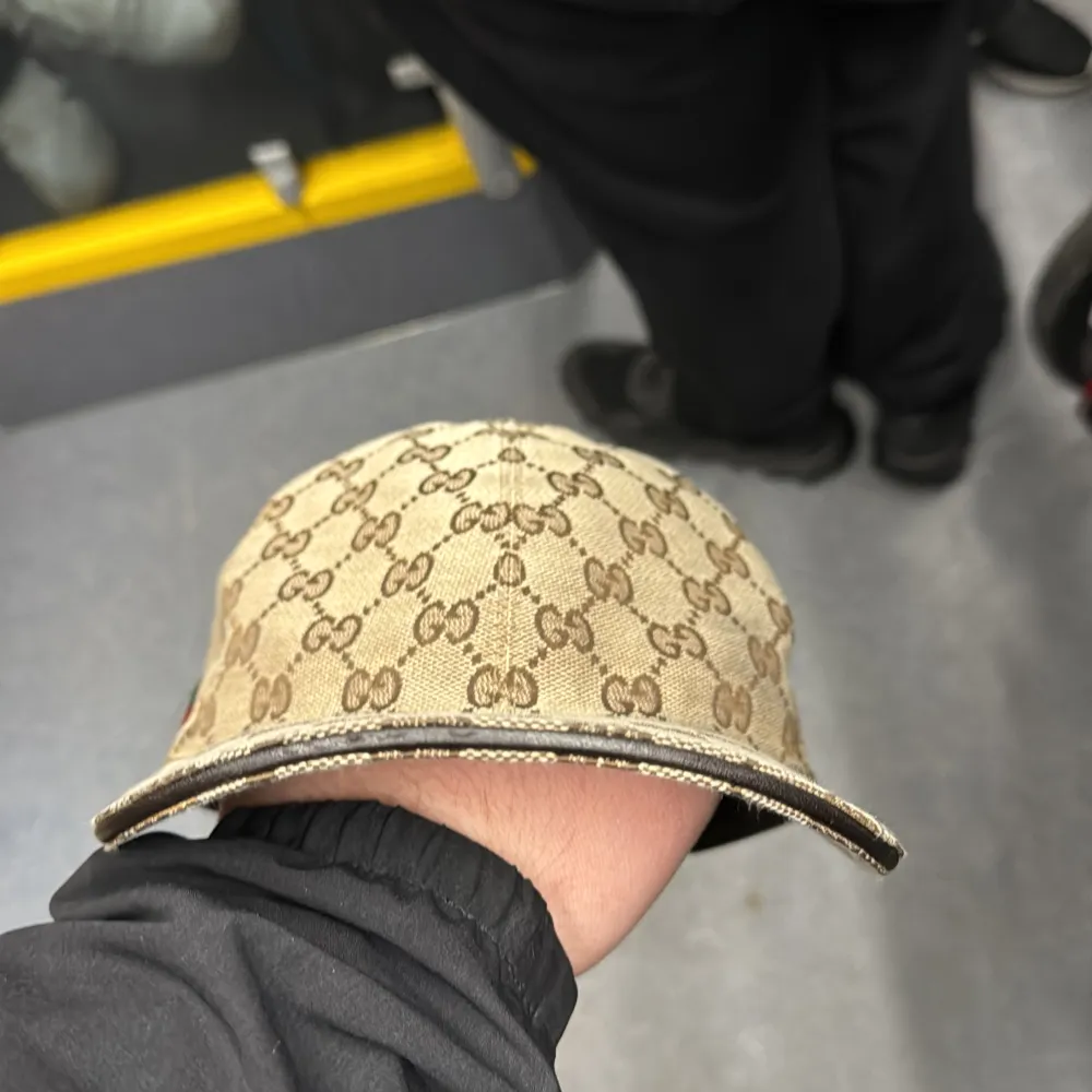 Säljer en snygg beige keps från Gucci med det klassiska GG-monogrammet. Kepsen har en justerbar rem baktill och en grön-röd randdetalj på sidan. Perfekt för att ge din outfit en lyxig touch!. Asusteet.