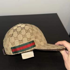 Snygg beige keps från Gucci med det klassiska monogrammönstret. Kepsen har en justerbar rem baktill och en grön och röd randdetalj på sidan. Perfekt för att ge din outfit en lyxig touch! Priset kan diskuteras. 