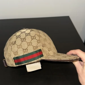 Beige Gucci keps - Snygg beige keps från Gucci med det klassiska monogrammönstret. Kepsen har en justerbar rem baktill och en grön och röd randdetalj på sidan. Perfekt för att ge din outfit en lyxig touch! Priset kan diskuteras. 