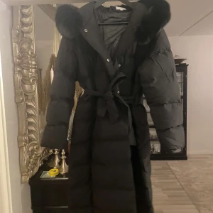 Svart dunjacka med pälshuva  - Säljer en svart lång dunjacka med pälshuva och knytbälte i midjan. Perfekt för kalla vinterdagar! Jackan är lång och varm samt har långa ärmar. Använd 1 gång, men den vart för stor för mig. Storlek L, säljer den för 1800 pris kan diskuteras🫶🏽